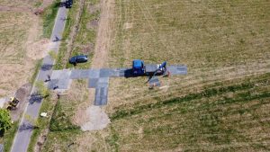 Pracownicy układają asfalt na nowo powstającej drodze w polu; w tle widoczny pojazd i maszyna budowlana.