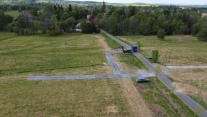 Widok z góry na wiejską okolicę z drogą, ciężarówką oraz pracownikami budowlanymi. W tle zieleń i domy.