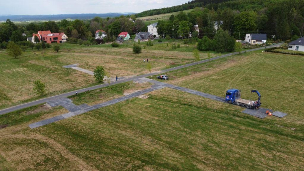 Widok na teren budowy z nowymi drogami, pojazdami i zielonymi polami, w tle widoczne domy i drzewa.