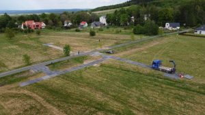 Widok na teren budowy z nowymi drogami, pojazdami i zielonymi polami, w tle widoczne domy i drzewa.