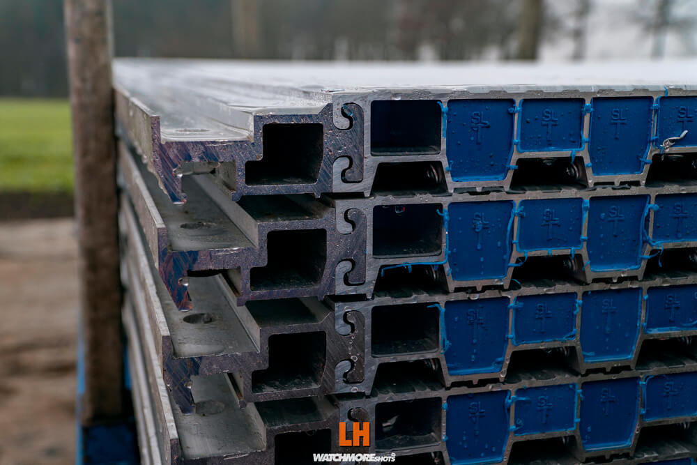 Stos aluminiowych profili, z widocznymi niebieskimi dolnymi krawędziami, ułożonych na zewnątrz.