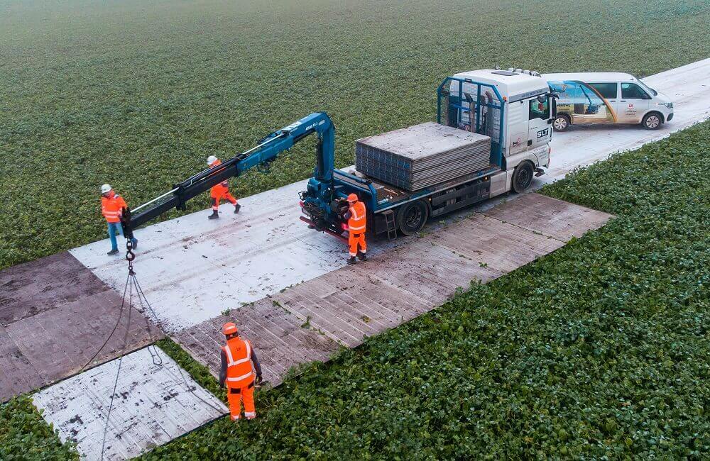 Pracownicy w pomarańczowych kamizelkach transportują płyty na polu z pojazdem ciężarowym. W tle roślinność.
