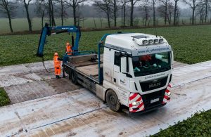 Ciężarówka MAN na polu, z robotnikami w pomarańczowych ubraniach ładującymi materiały przy użyciu dźwigu.
