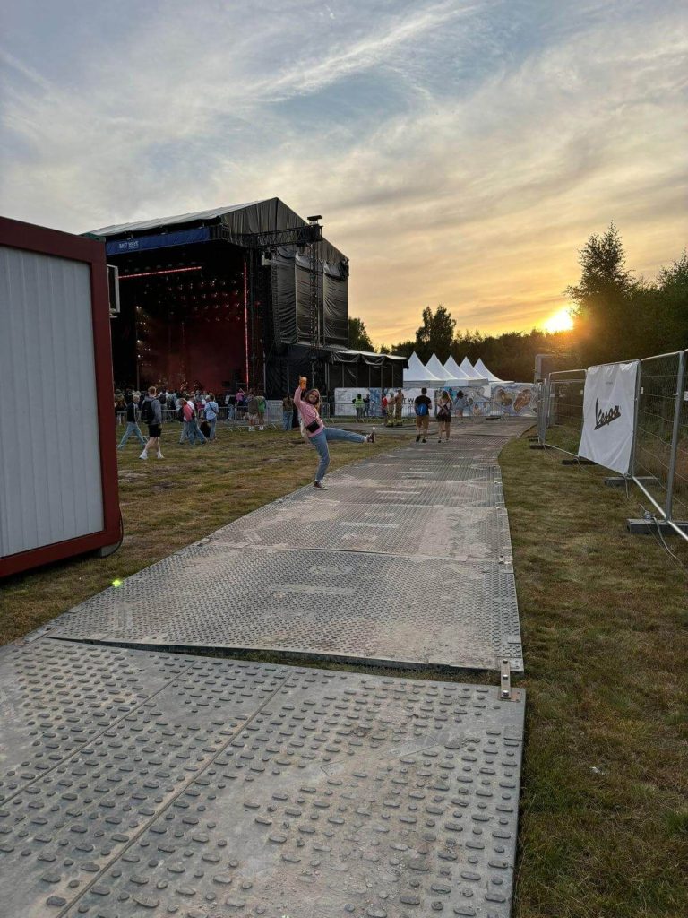 Na zdjęciu scena koncertowa z ludźmi, w tle zachód słońca i namioty. Widoczna droga z materiału metalowego.