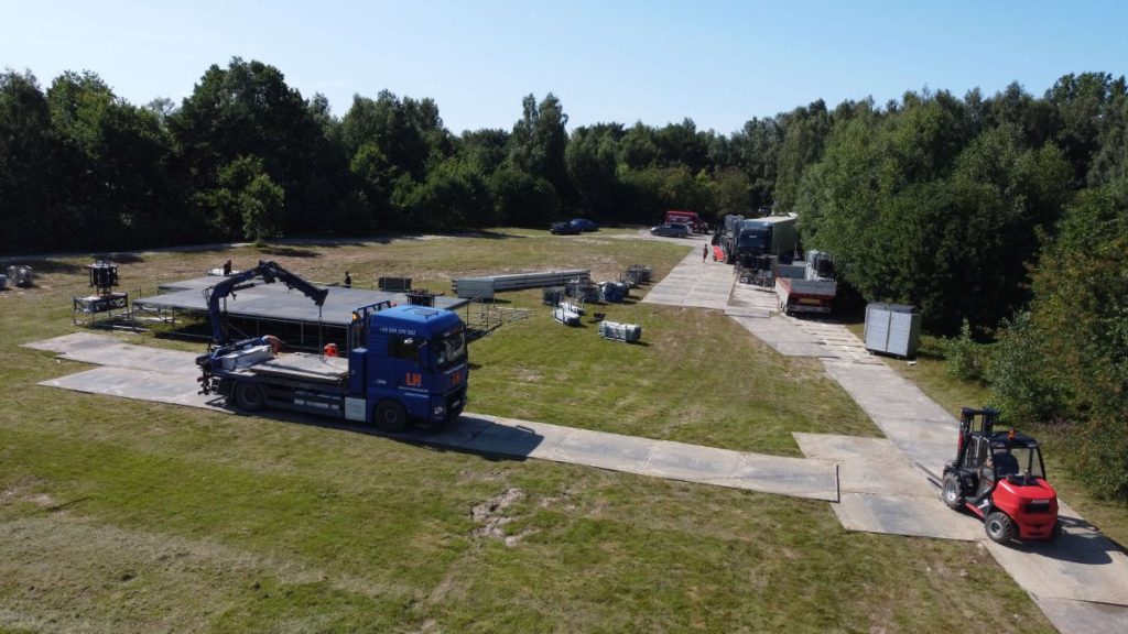 Widok na plac budowy z ciężarówką, wózkiem widłowym i materiałami budowlanymi na trawie w otoczeniu drzew.