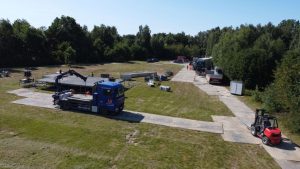 Widok na plac budowy z ciężarówką, wózkiem widłowym i materiałami budowlanymi na trawie w otoczeniu drzew.