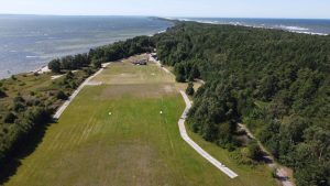 Widok na zieloną łąkę z plażą i lasem w tle, z morzem po lewej stronie. W oddali widać kilka obiektów.