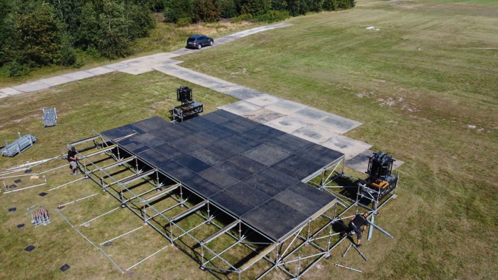 Widok na plac budowy sceny, z stojakami i sprzętem. W tle widać zieleń oraz samochód na utwardzonej nawierzchni.