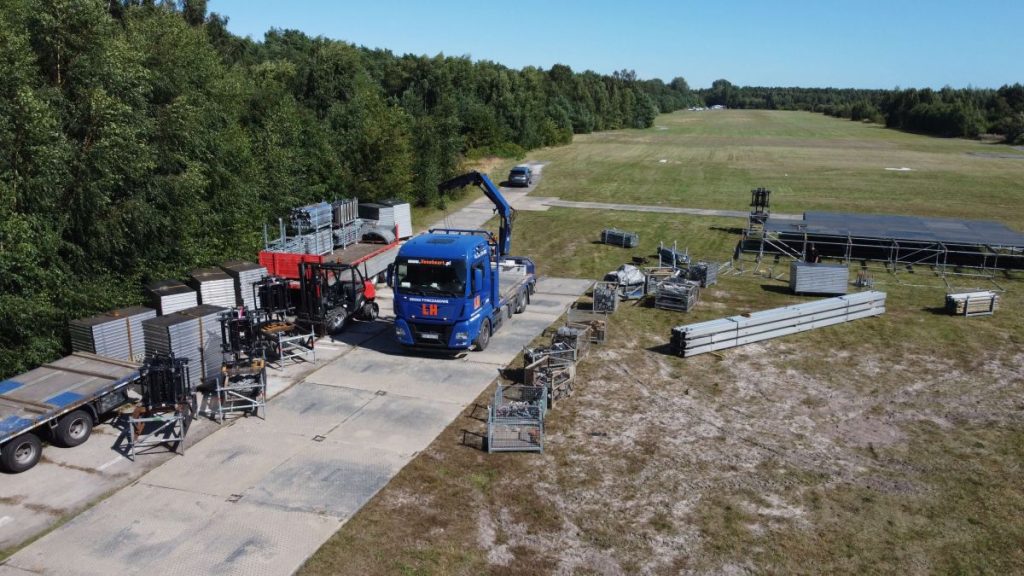 Zespół sprzętu budowlanego na placu, niebieski ciężarówka z dźwigiem oraz stogi materiałów na trawie.