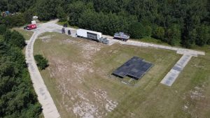 Pusty teren z ciężarówką i konstrukcją do występów, otoczony drzewami. W tle wyraźne ślady po drodze.