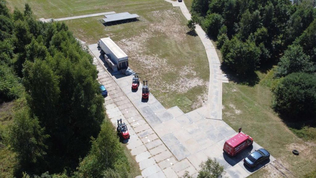 Widok z góry na teren z betonowymi nawierzchniami, wózkami widłowymi i pojazdami na otwartej przestrzeni otoczonej drzewami.