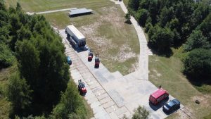 Widok z góry na teren z betonowymi nawierzchniami, wózkami widłowymi i pojazdami na otwartej przestrzeni otoczonej drzewami.