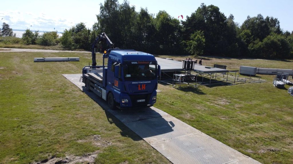 Ciężarówka na podjeździe z metalowymi konstrukcjami w tle, otoczona zielonym terenem i drzewami.