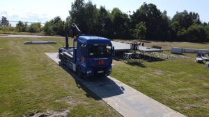 Ciężarówka na podjeździe z metalowymi konstrukcjami w tle, otoczona zielonym terenem i drzewami.