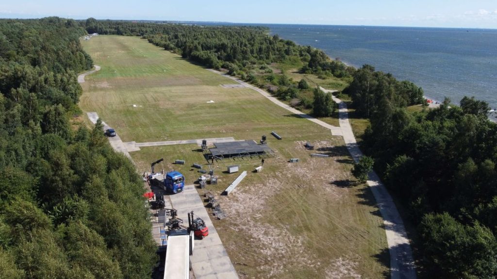 Widok na otwartą przestrzeń z grasszem, drogą, pojazdami oraz bliskim brzegiem wody. Zalesiony teren w tle.