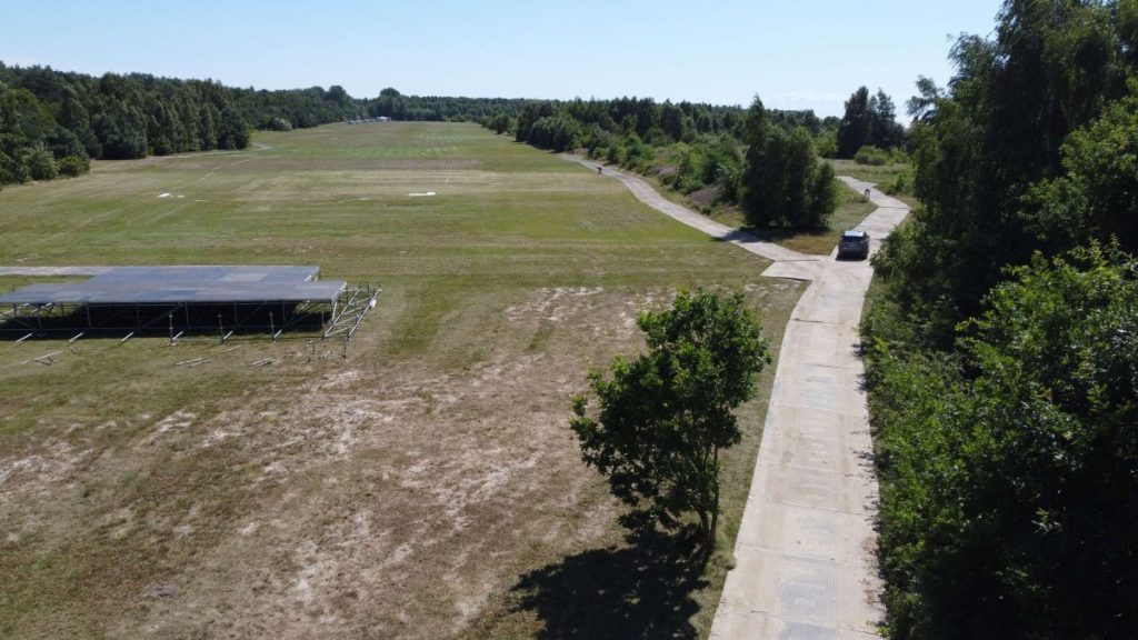 Widok na otwartą przestrzeń z trawnikiem, alejką i samochodem w oddali. W tle drzewa i pole.