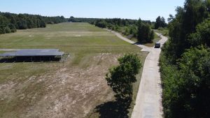 Widok na otwartą przestrzeń z trawnikiem, alejką i samochodem w oddali. W tle drzewa i pole.