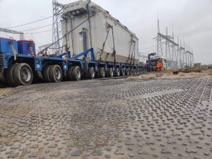 Transport dużego transformatora na naczepie, w pobliżu linii energetycznych, przy mokrej nawierzchni.