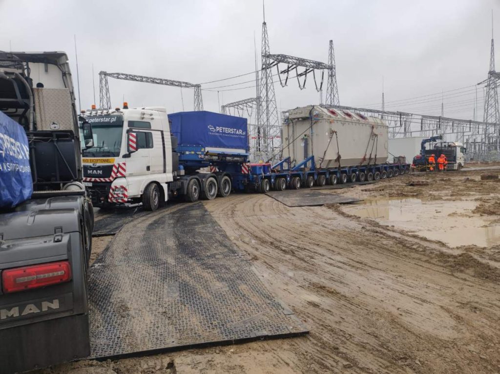 Ciężarówka transportująca duży ładunek przy stacji elektrycznej, otoczona pracownikami w odzieży roboczej.