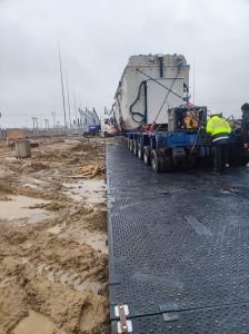Na obrazie widać ciężki sprzęt transportowany po tymczasowej drodze na placu budowy, otoczonej błotem.