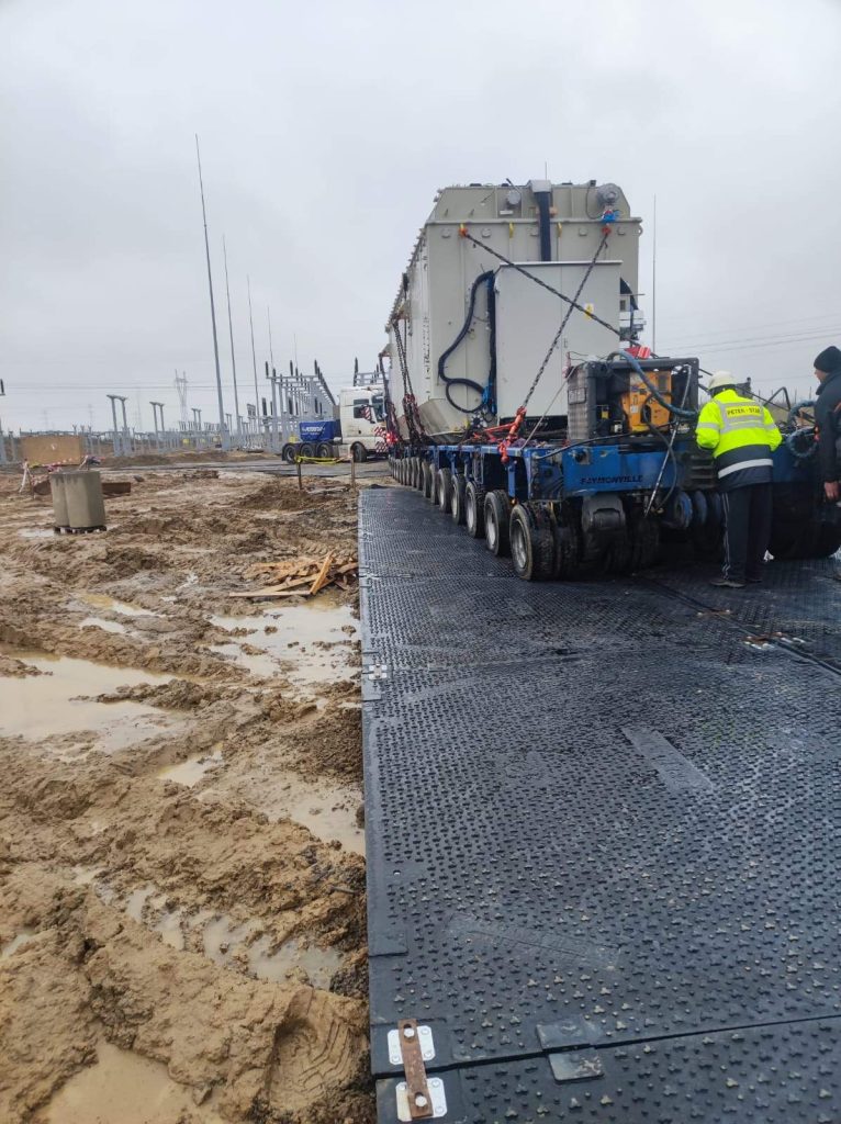 Na obrazie widać ciężki sprzęt transportowany po tymczasowej drodze na placu budowy, otoczonej błotem.