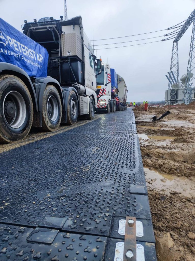 Ciężarówki na placu budowy, obok grząskiego terenu i linii energetycznych. Widoczna droga z płyty zabezpieczającej.