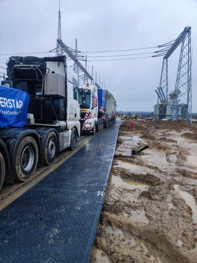 Zespół ciężarówek na plaży budowlanej, obok linii energetycznych, w otoczeniu błota i placu budowy.