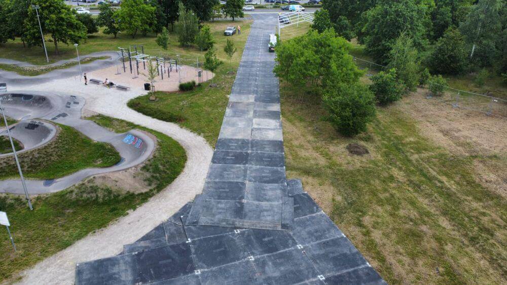 Ścieżka w parku z asfaltowym podłożem, obok strefa do ćwiczeń oraz tereny zielone.