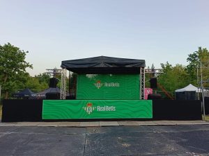 Scena z czarnym zadaszeniem, zielonym tłem z logo RealBetis, ustawiona na terenie z drzewami w tle.