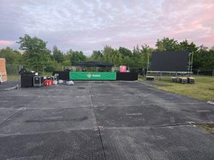 Przestrzeń koncertowa z czarną sceną, ekranem, sprzętem nagłośnieniowym i zielonym tłem, otoczona drzewami.