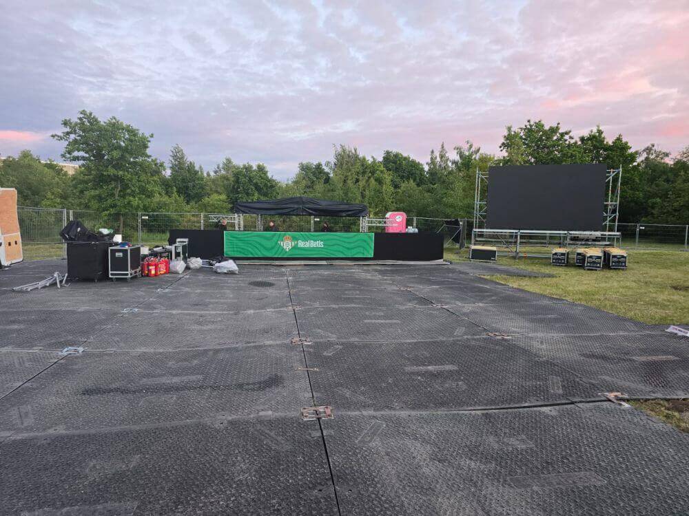 Przestrzeń koncertowa z czarną sceną, ekranem, sprzętem nagłośnieniowym i zielonym tłem, otoczona drzewami.