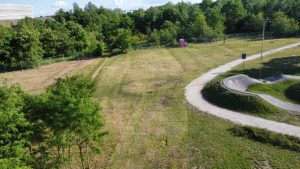 Widok z góry na otwartą przestrzeń z trawą, drzewami oraz ścieżką i placem zabaw. W tle widoczna toaleta przenośna.