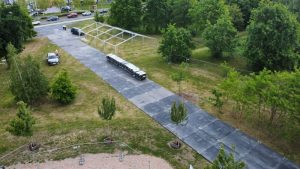 Widok z góry na aleję w parku, z pojazdem i ustawionymi kontenerami na odpady w tle, otoczone drzewami.