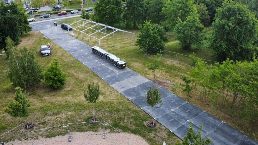 Widok z góry na aleję w parku, z pojazdem i ustawionymi kontenerami na odpady w tle, otoczone drzewami.