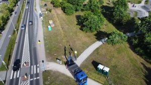 Widok z drona na drogę, z samochodem ciężarowym, zieloną trawą i alejkami w parku. W tle znaki i toalety przenośne.