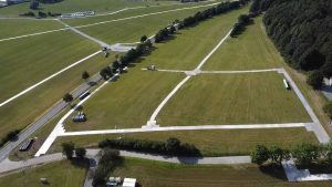 Widok na dużą, otwartą przestrzeń z wyznaczonymi ścieżkami i zielonymi terenami, otoczoną drzewami.