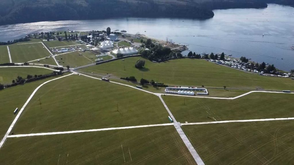 Widok z lotu ptaka na zielone pola, budynki i wodę w tle, tworzący malowniczy krajobraz nadbrzeżny.