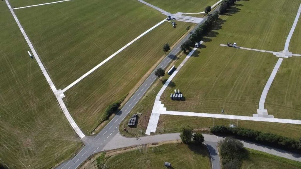 Widok z lotu ptaka na pola, drogę i kilka pojazdów na wyznaczonej przestrzeni. Równomiernie przycięta trawa.