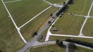 Widok z lotu ptaka na pola, drogę i kilka pojazdów na wyznaczonej przestrzeni. Równomiernie przycięta trawa.