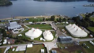 Widok na teren festiwalu z namiotami, wodą w tle oraz różnymi strukturami i pojazdami.