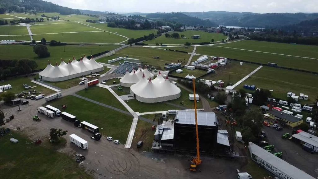 Widok z lotu ptaka na teren festiwalu z białymi namiotami i sceną, otoczony zielonymi wzgórzami.