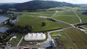 Widok z lotu ptaka na tereny zielone z namiotem, drogami i jeziorem w tle. W okolicy obozowisko i konstrukcje.