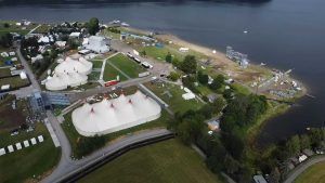 Widok lotniczy na teren festiwalu z białymi namiotami, wodą i zabudowaniami w tle.