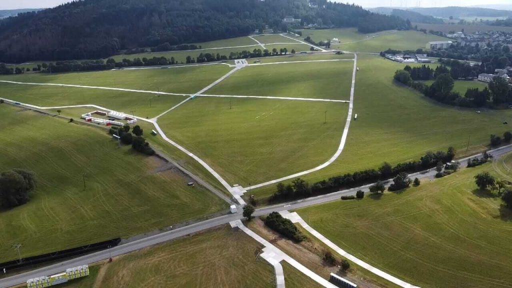 Widok z góry na rozległe zielone pole, drogi oraz zabudowania w tle, otoczone przyrodą i wzgórzami.