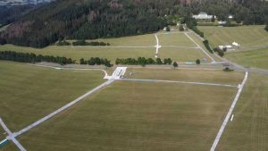 Widok lotniska z lotu ptaka, z dużymi pasami trawiastymi, drzewami w tle i budynkami na obrzeżach.