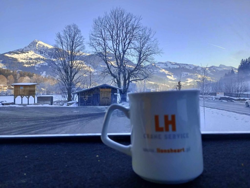 Kubek z napisem LH SERWIS na tle górskiego krajobrazu zimowego, z drzewami i śniegiem.