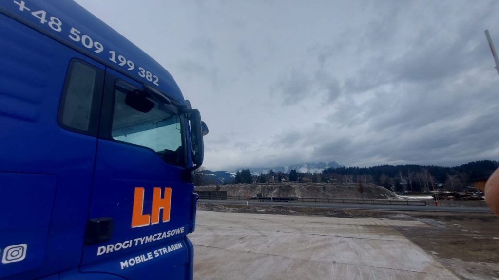 Niebieska ciężarówka z logo LH, w tle góry i pochmurne niebo.
