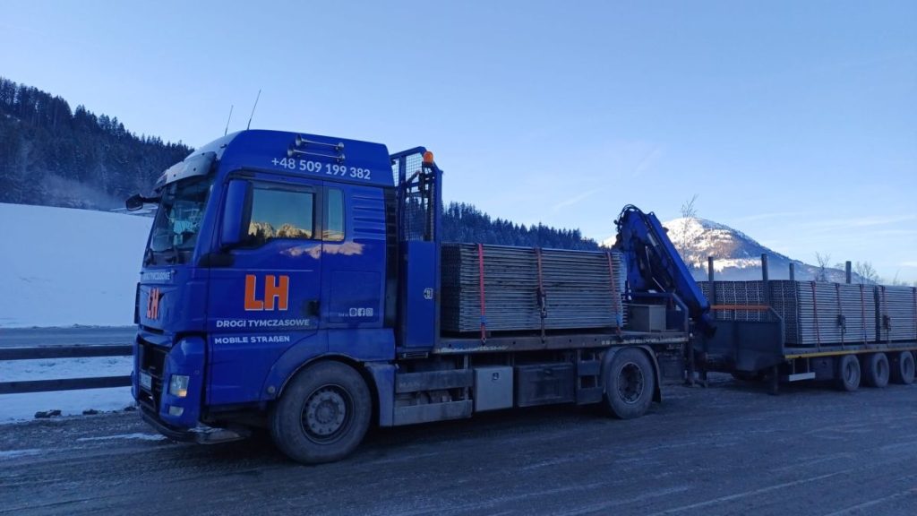 Niebieski ciężarówka z długim przyczepem, załadowana drewnem, na tle górskiego krajobrazu i jasnego nieba.