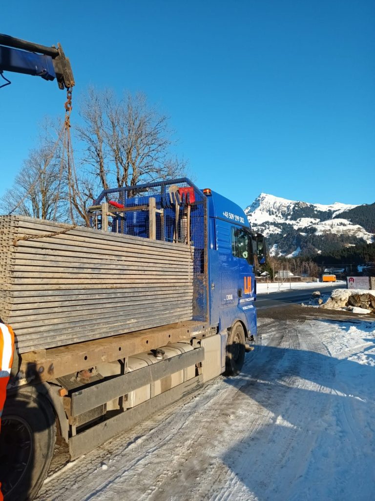 Niebieski ciężarówka z platformą, zaśnieżona droga i góry w tle.