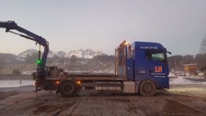 Ciężarówka z niebieską kabiną i długą przyczepą, z widokiem na górskie szczyty w tle, w zimowej scenerii.